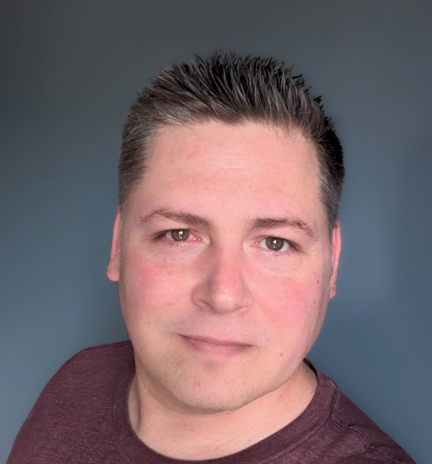 Brad Tetlow The image shows a man with short, neatly styled dark hair. He has fair skin and is wearing a maroon shirt. He is looking directly at the camera with a neutral expression and a slight smile. The background is a solid, muted gray-blue color, keeping the focus on his face.