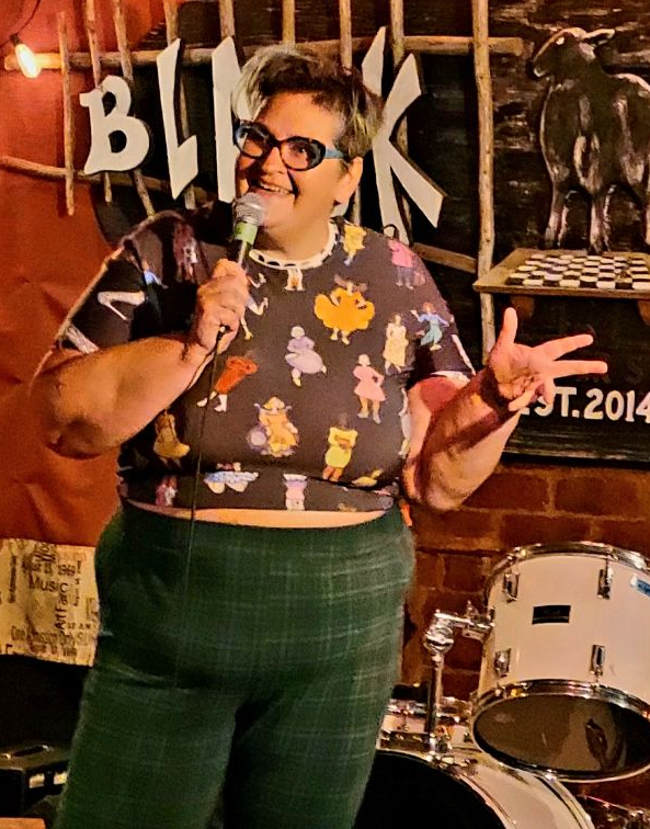 Cara Winsor Hehir The image shows a woman performing stand-up comedy on a stage. She has short hair and is wearing glasses. Her outfit includes a colorful patterned top with various cartoon characters and high-waisted green plaid pants. She is holding a microphone with one hand and making a peace sign with the other, smiling and engaging with the audience. The background features a brick wall with string lights and musical instruments, including a drum set, creating a cozy and lively atmosphere.