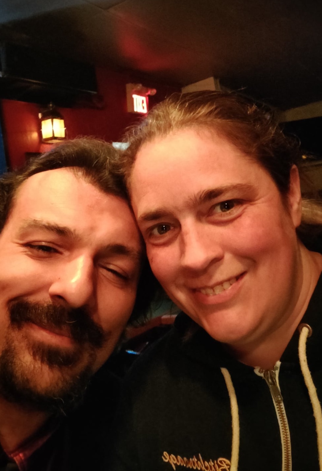 Chris Lansdell and Sarah Macfie The image is a close-up selfie of a man and a woman. They are smiling and appear to be in a cozy, dimly lit indoor setting. The man has short dark hair and a beard, and his eyes are partially closed as he smiles. The woman has light brown hair pulled back and is wearing a hoodie. The background includes warm lighting and an exit sign, adding to the intimate, casual atmosphere.