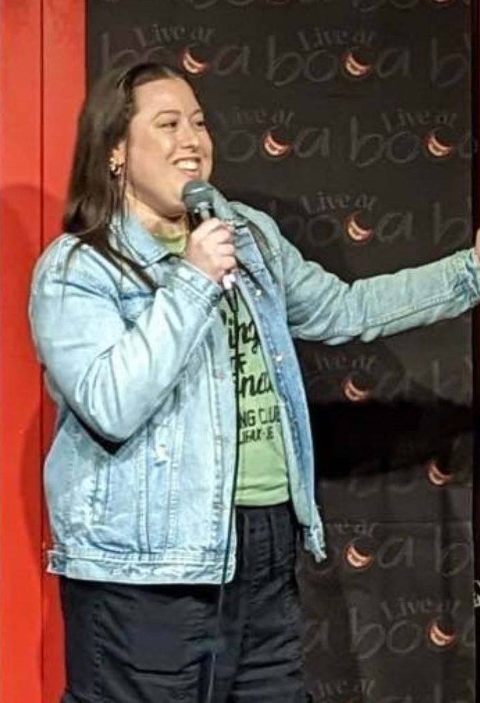 Jenna Makrides The image shows a woman performing stand-up comedy on stage. She has long dark hair and is wearing a light denim jacket over a green shirt, black cargo pants, and white sneakers. She is holding a microphone with one hand and the mic stand with the other, smiling as she engages with the audience. The backdrop features the logo "Live at Boa" and some lighting, creating a cozy comedy club atmosphere. The stage area has a red door and wooden floor, adding warmth to the setting.