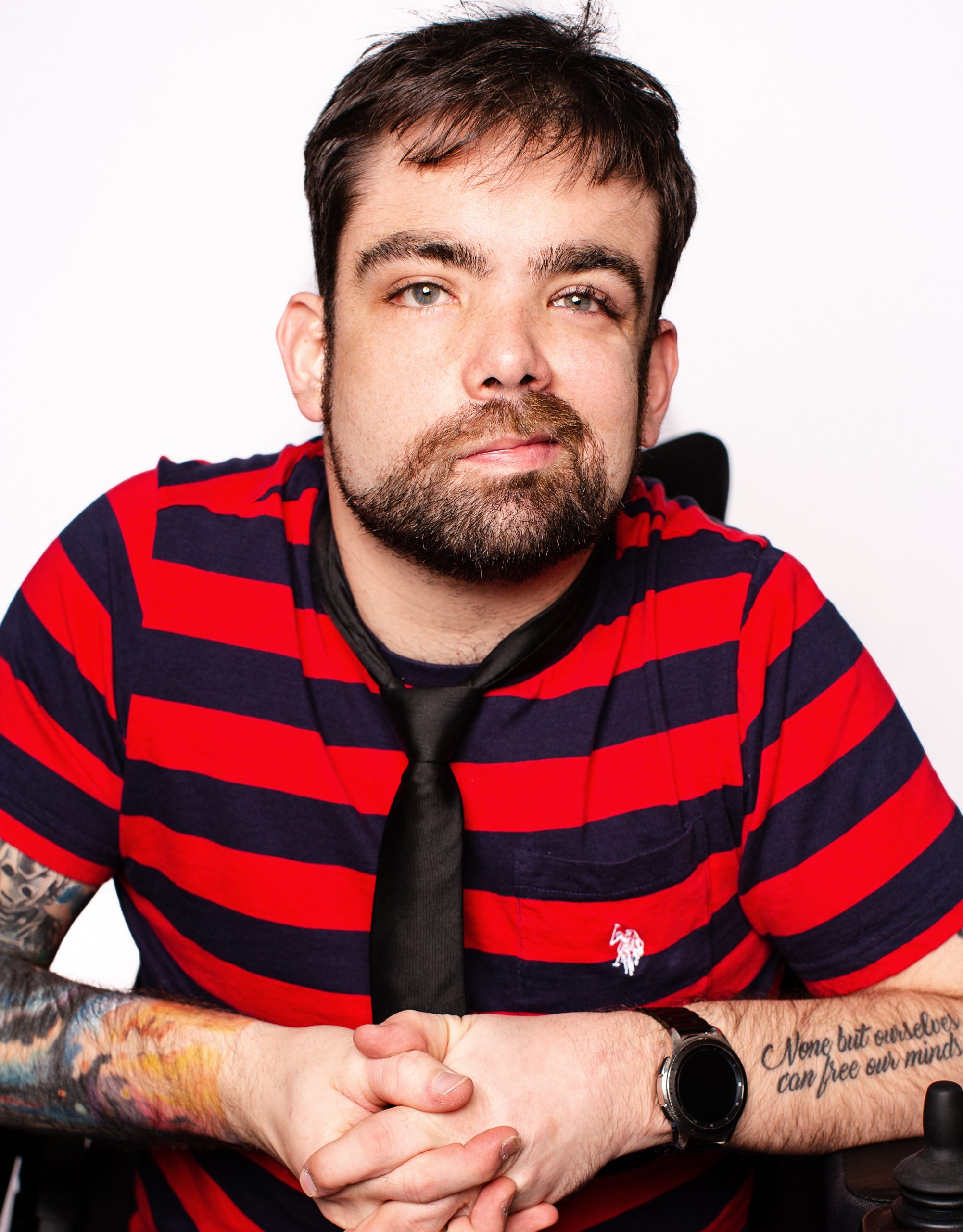 Josh Menchions The image shows a man with short dark hair and a beard, wearing a red and navy striped shirt, a black necktie, and jeans. He is sitting and has his hands clasped in front of him. He has tattoos on his arms, one of which has text that reads, "None but ourselves can free our minds." He is looking directly at the camera with a calm, thoughtful expression. The background is plain and white, keeping the focus on the man.