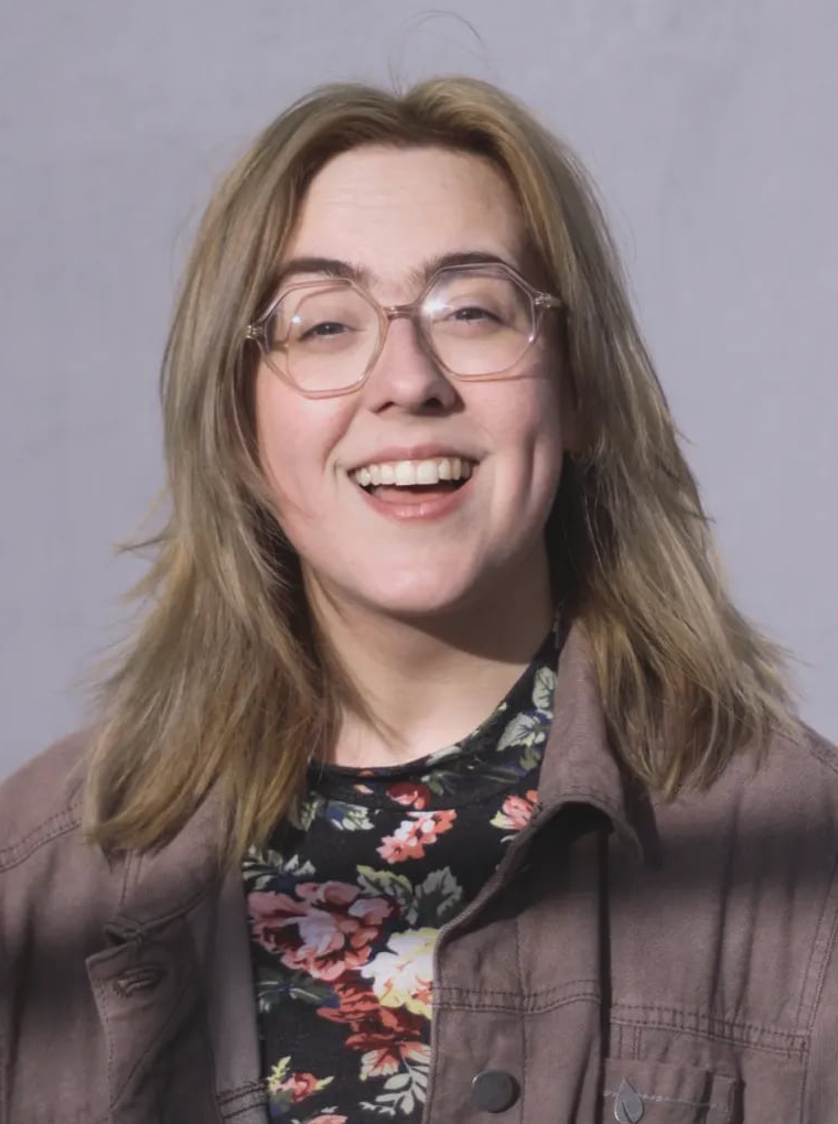 Katy Warren The image shows a smiling woman with light brown hair that falls just below her shoulders. She is wearing glasses and a floral-patterned top underneath a brown jacket. The background is plain and gray, focusing attention on her happy and approachable expression.