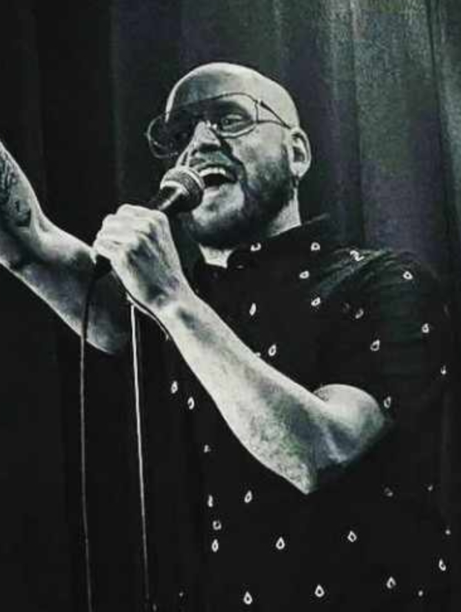 Liam Small The image shows a man performing stand-up comedy on stage in a black and white setting. He is bald and has a beard, wearing glasses and a patterned short-sleeved shirt. He is holding a microphone with one hand and pointing upwards with the other, engaging energetically with the audience. The background is dark, enhancing the focus on the performer. His stance and expression suggest he is in the middle of delivering a punchline or an animated part of his act.
