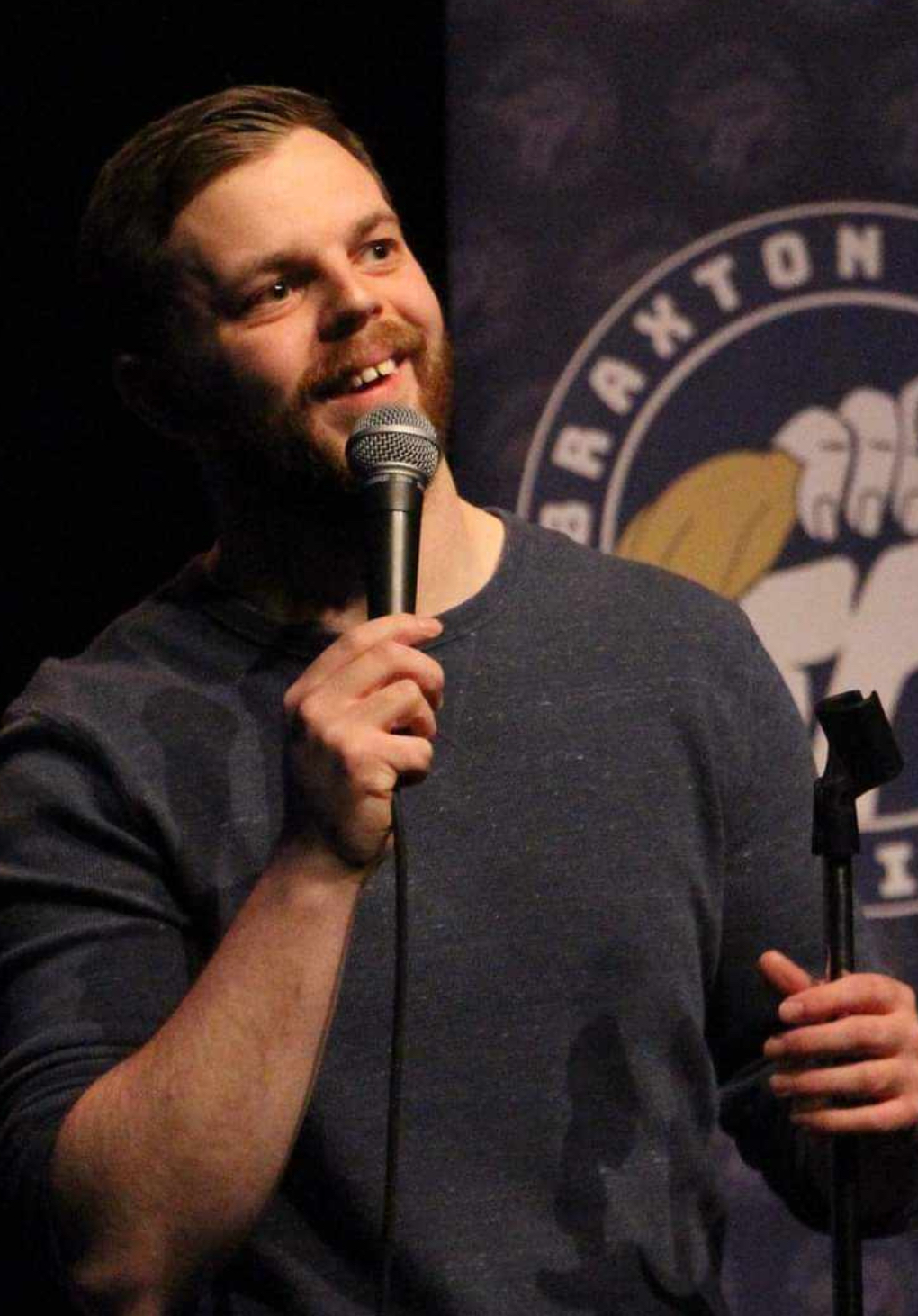 Stephen Vanier The image shows a man performing stand-up comedy on stage. He has short hair and a beard and is wearing a dark long-sleeve shirt. He is holding a microphone and smiling, looking engaged with the audience. Behind him is a banner with the logo of the "Braxton Comedy Festival," featuring an illustration of a fist holding a yellow chicken. The background is dark, highlighting the performer and the banner. The overall atmosphere suggests he is in the middle of a lively and enjoyable performance