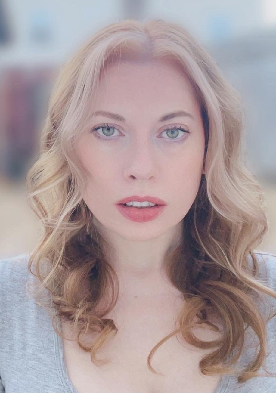 Susan Kelsey The image shows a woman with long, wavy blonde hair. She is wearing a gray short-sleeved top and has a neutral expression on her face. Her eyes are light-colored, and she has a calm and focused gaze. The background is blurred, featuring outdoor elements such as a fence and buildings, suggesting she is outside. The overall atmosphere is serene and natural.