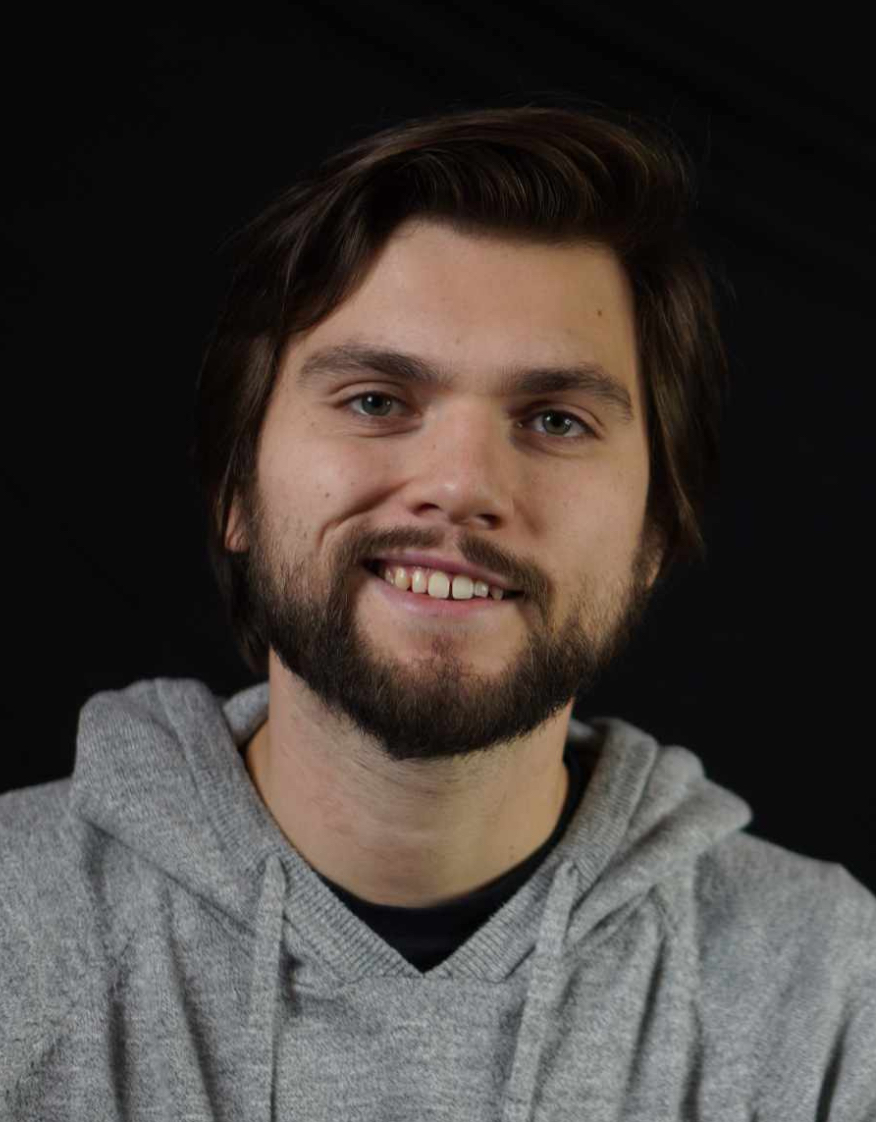 Todd Butt The image shows a man with short dark hair and a beard, smiling at the camera. He is wearing a light gray hoodie. The background is plain black, keeping the focus on his face. His expression is friendly and approachable.