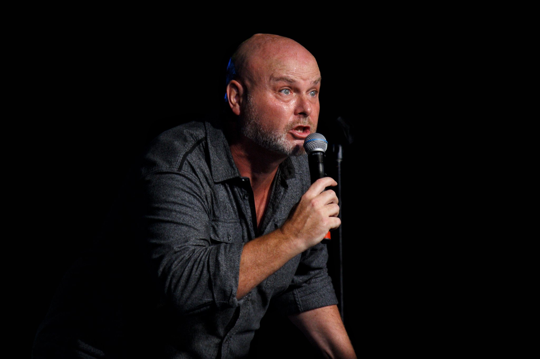 411536634_891905669604774_3968534617428463729_n The image shows a bald man with a beard performing stand-up comedy on stage. He is holding a microphone in one hand and appears to be in the middle of delivering a joke or story, with an animated expression on his face. He is wearing a dark, long-sleeve shirt, and the background is completely black, highlighting him as the focal point of the image. The lighting emphasizes his features and creates a dramatic, engaging atmosphere typical of a comedy performance.