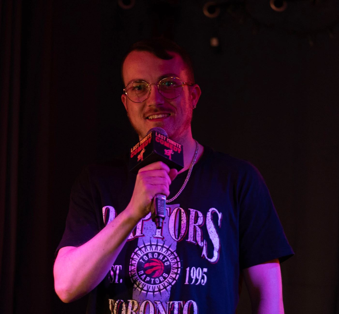 Aaron Andrews Subject Person: A man with glasses and short hair. Appearance Clothing: He is wearing a black T-shirt with "Toronto Raptors" and the Raptors logo printed on it. Facial Features: He has short hair and is wearing round glasses. Expression: He is smiling. Accessories: He is holding a microphone with a "Last Minute Comedy" label on it, suggesting he is performing or speaking. Background Lighting: The background is dark, and the lighting highlights the person. Additional Elements: There are no additional elements visible in the background, keeping the focus on the person. This image captures a moment of a man enjoying his time on stage, with the microphone and lighting suggesting a performance or comedic act.