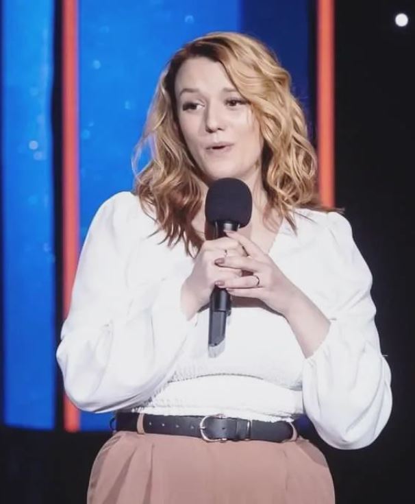 Bree Parsons The image shows a woman with shoulder-length, wavy blonde hair performing stand-up comedy. She is holding a microphone with both hands and has a friendly, engaging expression. She is wearing a white blouse with long sleeves and a pair of high-waisted, light brown pants with a black belt. The background features vertical colored lights, creating a vibrant and lively stage atmosphere.
