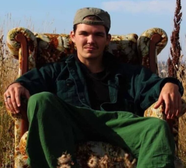 Christian Leonard The image shows a young man sitting in an old-fashioned, upholstered armchair placed outdoors in a field. He is wearing a green jacket, matching green pants, and a backwards baseball cap. His expression is relaxed and confident as he leans back in the chair with his hands resting on the armrests. The setting features tall grasses and wildflowers, creating a natural, serene atmosphere under a clear blue sky.
