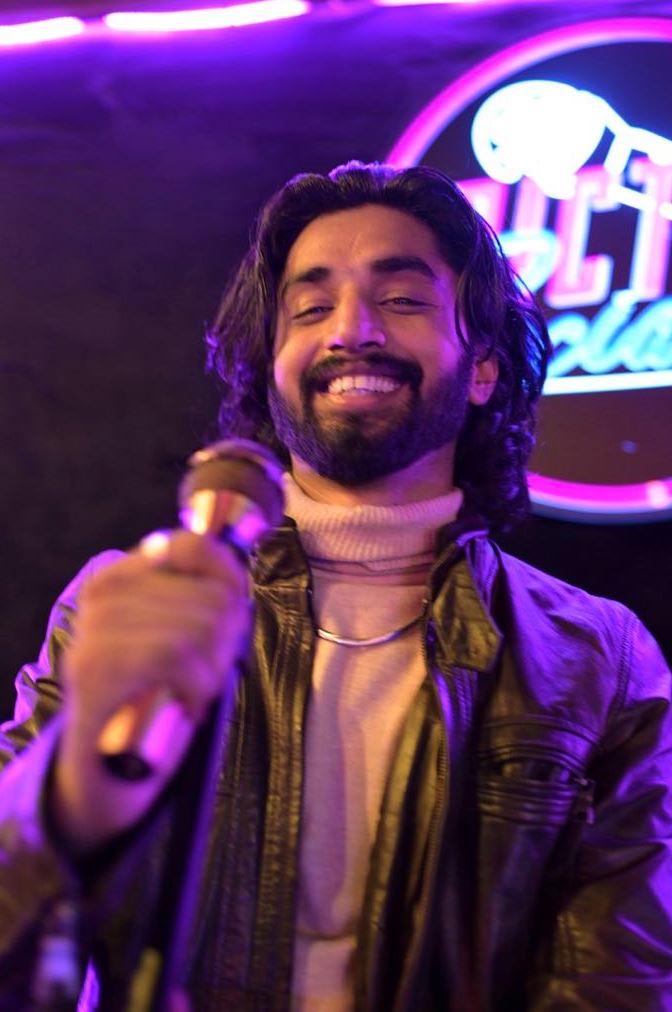 Haroon Malik Subject Person: A man with long hair and a beard. Appearance Clothing: He is wearing a light-colored turtleneck and a leather jacket. Facial Features: He has long, dark hair and a beard. Expression: He is smiling warmly. Accessories: He is holding a microphone, suggesting he is performing or speaking. Background Lighting: The background is dark with purple lighting, creating a vibrant and dynamic atmosphere. Additional Elements: There is a neon sign in the background, adding to the lively setting. This image captures a moment of a man enjoying his time on stage, with the colorful lighting and neon sign enhancing the energetic ambiance.