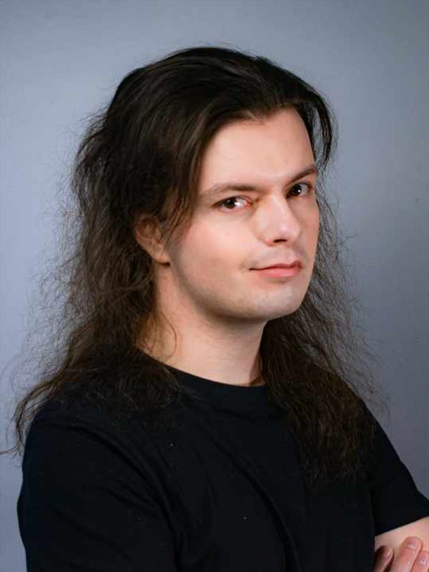 Jarod Farrell The image shows a man with long, dark, slightly wavy hair and fair skin. He is wearing a black shirt and has a subtle smile on his face. He is looking directly at the camera with a calm and composed expression. The background is plain and gray, keeping the focus on his face and upper body.