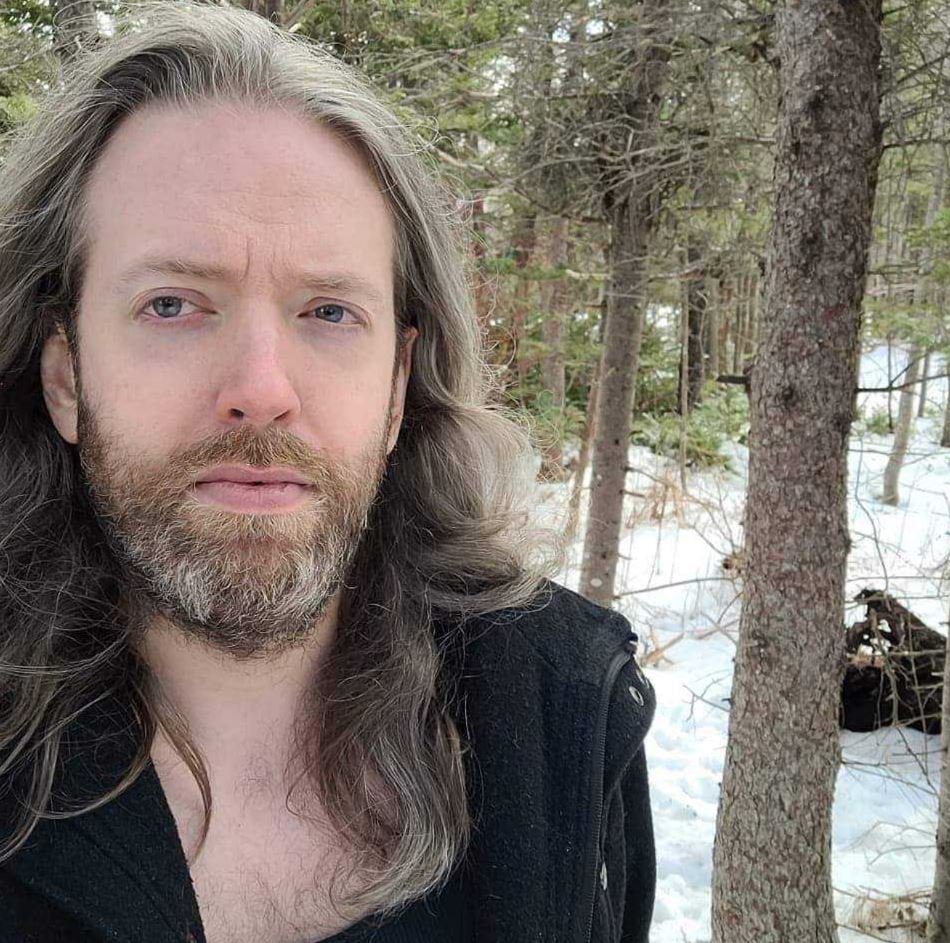 Josh Taylor Subject Person: The person in the image appears to be a male with long hair and a beard. Appearance Hair: Long, grayish hair. Clothing: Wearing a dark jacket or coat. Expression: The expression is neutral, possibly slightly contemplative, with direct eye contact with the camera. Background Setting: The background suggests an outdoor environment, possibly a forest or wooded area. There is snow on the ground and trees in the background. This image captures a serene and natural moment, with the subject standing outdoors in a wintery landscape, providing a calm and thoughtful atmosphere.