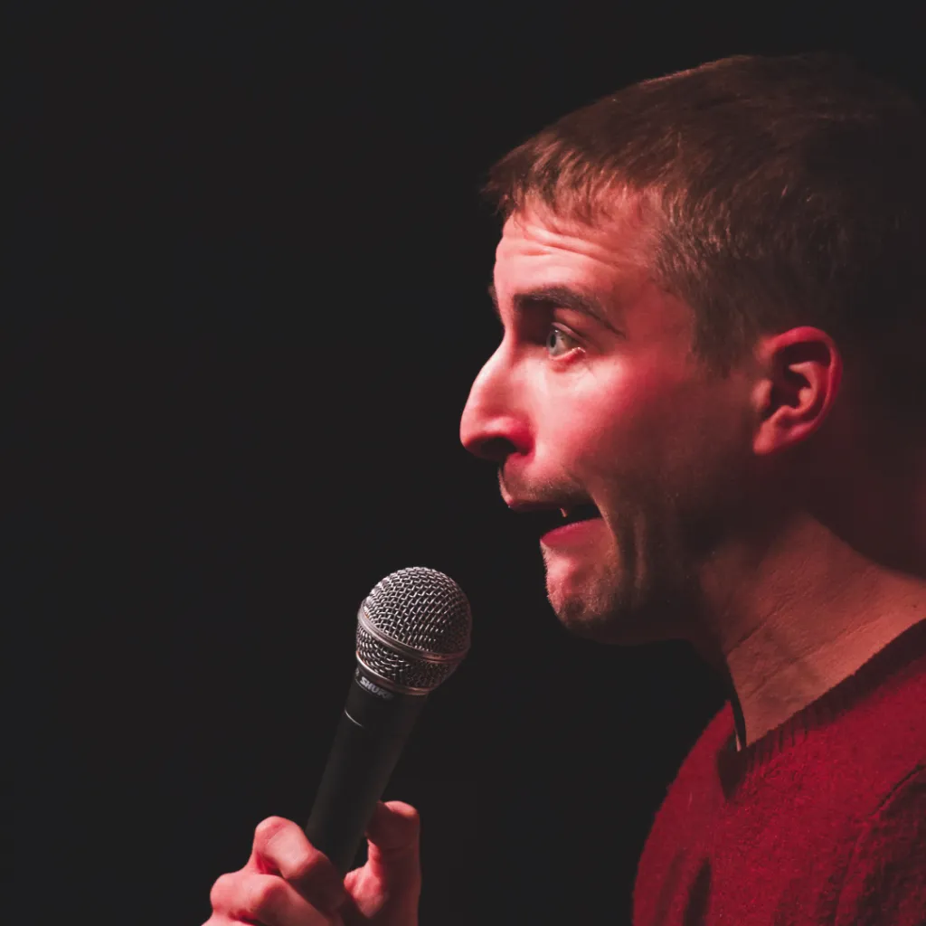 Mike- The image shows a man with short dark hair performing stand-up comedy on stage. He is holding a microphone close to his mouth with both hands and has an expressive, animated facial expression, suggesting he is in the middle of delivering a joke or a punchline. He is wearing a dark-colored top. The background is dark, focusing attention on the performer and his expressive demeanor.