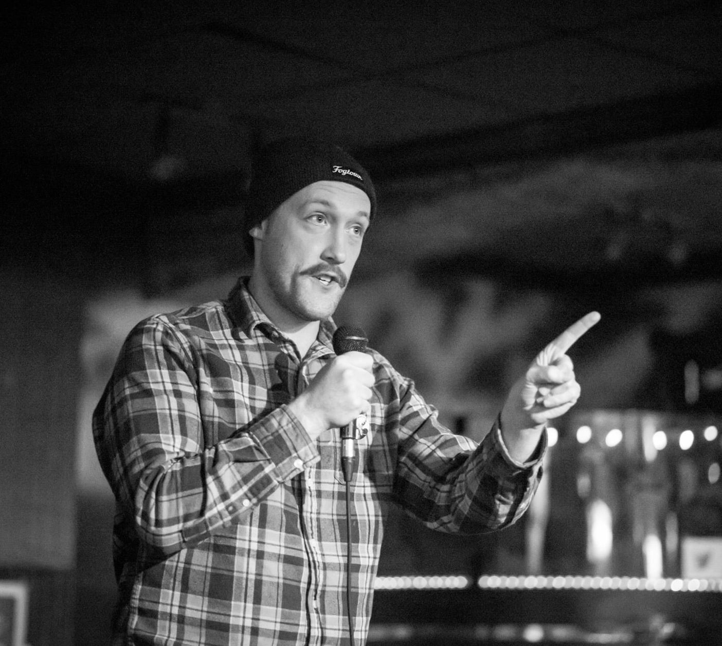 Mike Fardy Subject Person: A man performing stand-up comedy. Appearance Clothing: He is wearing a plaid shirt and a beanie hat. Facial Expression: He has a focused and engaged expression, interacting with the audience. Accessories: He is holding a microphone in one hand. Environment Background: The background appears to be a dimly lit bar or club setting, creating an intimate atmosphere. Stage Setup: There are lights in the background, possibly from the bar or stage setup, creating a warm and inviting ambiance. Lighting: The lighting is focused on the performer, highlighting his presence on the stage.