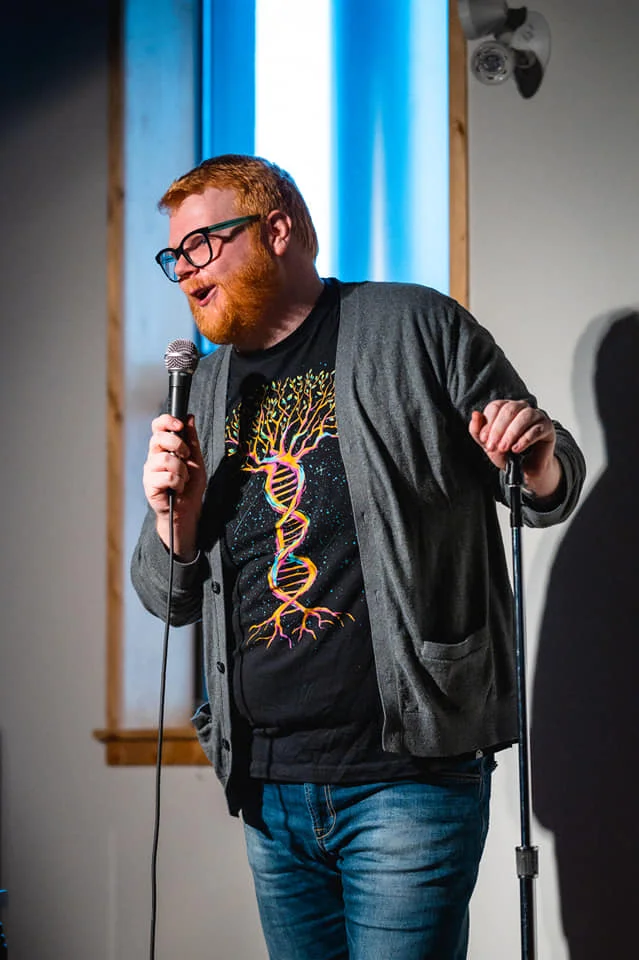 Mike Hammond The image shows a man with red hair and a beard performing stand-up comedy. He is wearing glasses, a gray cardigan, and a black T-shirt with a colorful DNA double helix design that transitions into tree roots. He is holding a microphone in one hand and the stand in the other, smiling and speaking, suggesting he is in the middle of a joke or story. The background features a lit window, creating a bright and engaging setting. The overall atmosphere is lively and engaging, characteristic of a comedy performance.