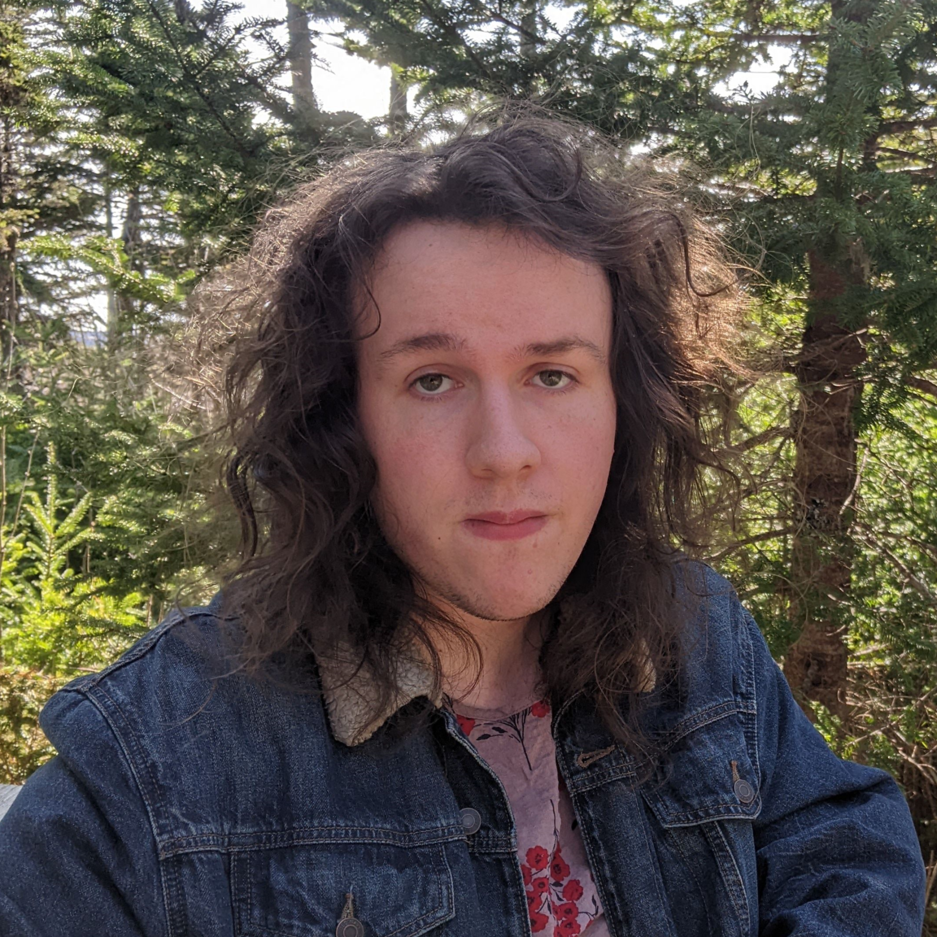 Nick Gushue Headshot 2023 The image shows a young person with long, wavy dark hair. They are wearing a denim jacket with a shearling collar over a floral-patterned top. Their expression is calm and relaxed, looking directly at the camera. The background features lush green trees, suggesting the photo was taken outdoors in a forest or park setting. The overall atmosphere is serene and natural.