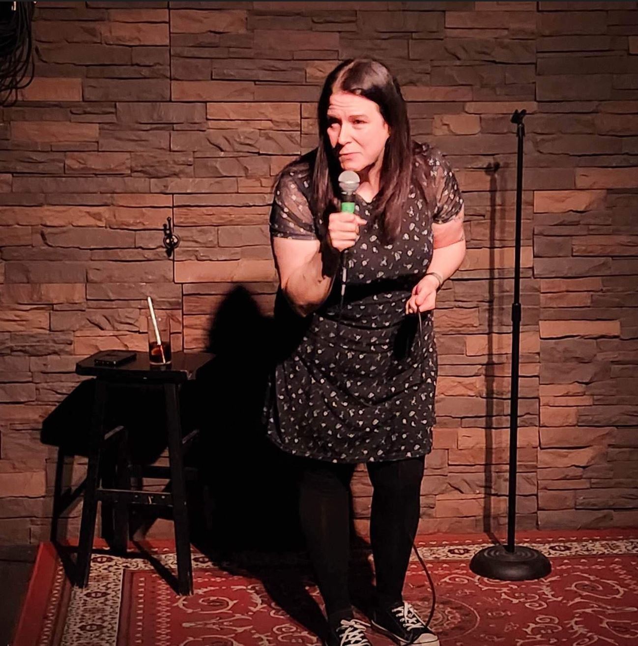 Nicole Downton Subject Person: A woman performing stand-up comedy. Appearance Clothing: She is wearing a dark dress with a floral pattern, black tights, and black sneakers. Facial Expression: She has a focused and animated expression, engaging with the audience. Accessories: She is holding a microphone in one hand. Environment Background: The backdrop consists of a brick or stone wall, giving a rustic and intimate vibe to the setting. Stage Setup: There is a small table or stool next to her with a drink and some items on it. A microphone stand is positioned behind her. The stage has a red patterned carpet. Lighting: The stage lighting is focused on her, highlighting her presence and creating a shadow against the wall. This image captures a moment of live comedy, showcasing the performer in an intimate and cozy setting, with an emphasis on her engaging stage presence.