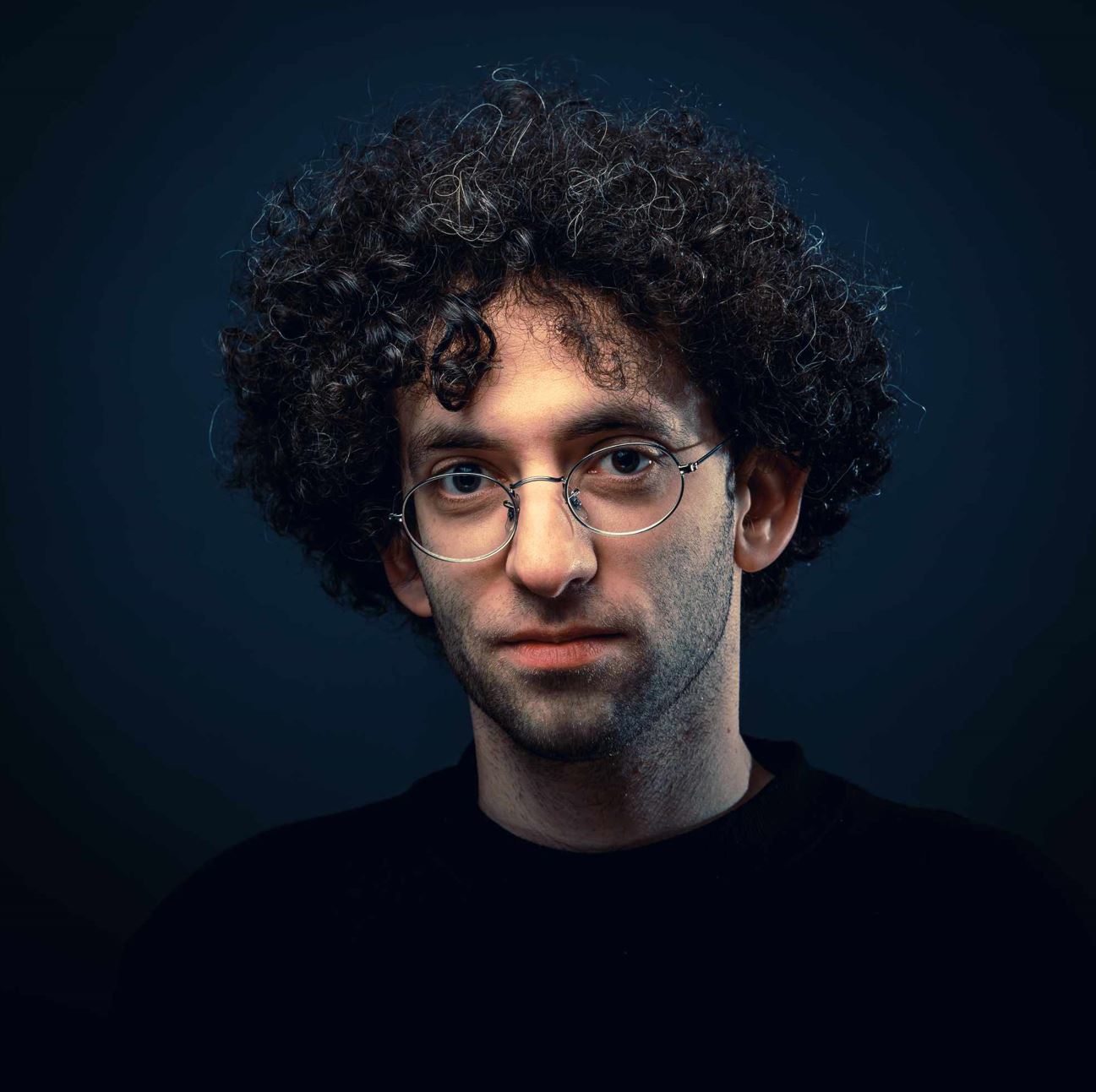 Paul Warford Subject Person: A man with curly hair and glasses. Appearance Clothing: He is wearing a dark top. Facial Features: He has distinct curly hair and is wearing round glasses. Expression: His expression appears calm and neutral. Background Lighting: The background is dark, which creates a contrast that highlights the subject's face and hair. This image captures a close-up portrait of the man, emphasizing his facial features and the texture of his curly hair. The dark background and lighting focus the viewer's attention on the subject.