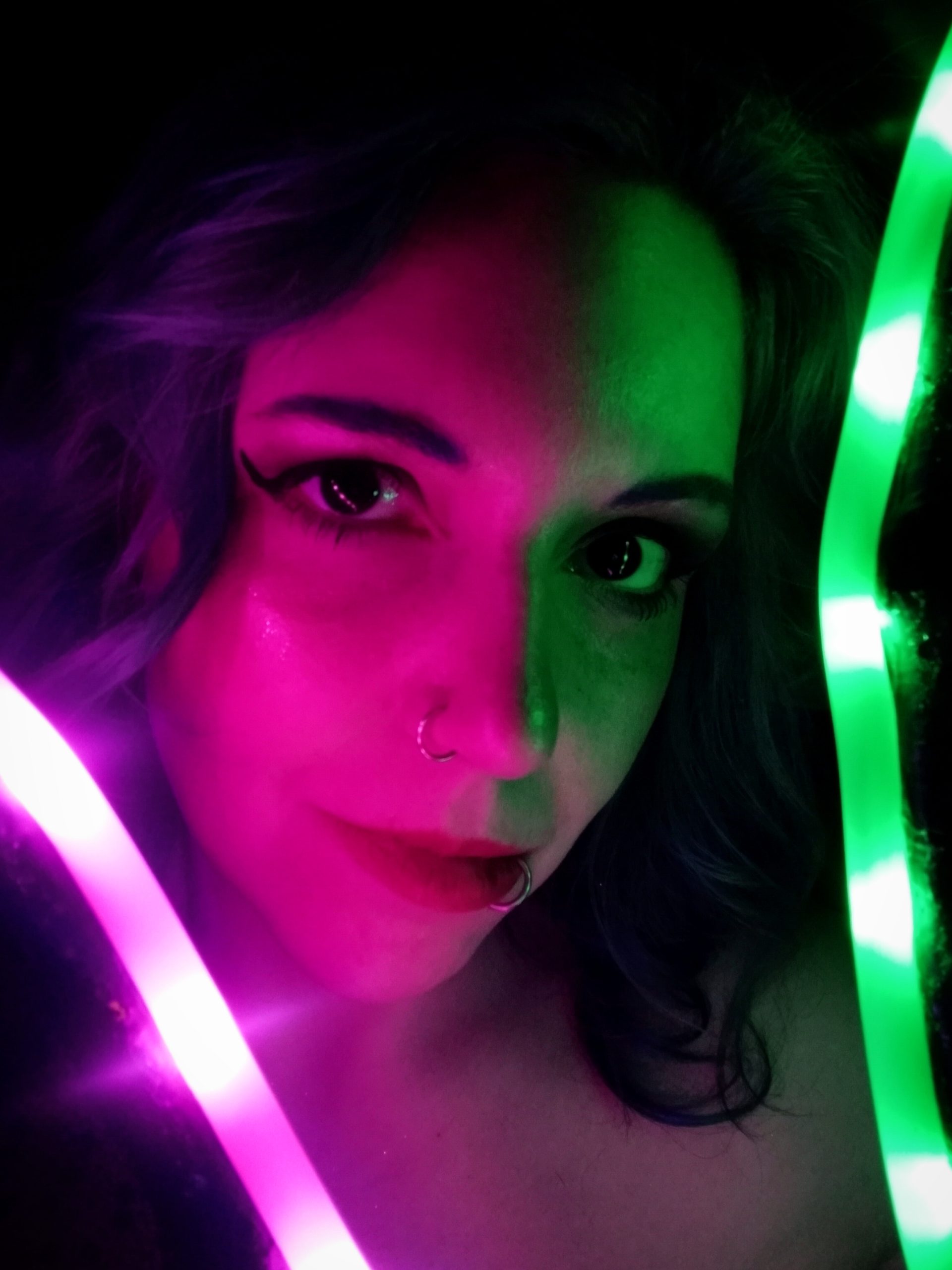 Tabitha Katz Subject Person: A woman with distinctive makeup and piercings. Appearance Facial Features: She has bold eyeliner, a nose ring, and a lip ring. Lighting: The image is creatively lit with neon lights, casting green and pink hues across her face. Expression: She has a calm and confident expression, looking directly at the camera. Background Lighting: The background is dark, making the neon lights and her features stand out more dramatically. Additional Elements: There are bright neon lights, one pink and one green, framing her face and adding to the artistic effect of the photograph. This image captures a striking and artistic portrait, with the neon lights providing a unique and vibrant contrast to the subject's features.