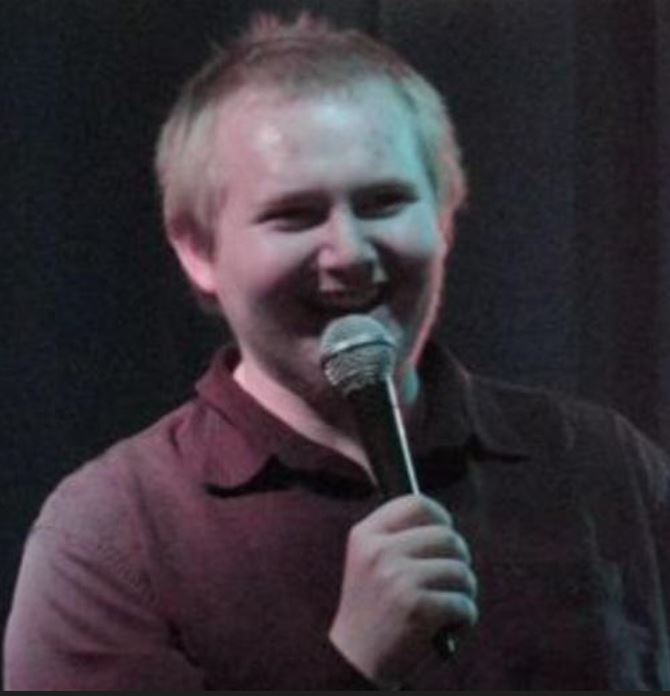 Tanner Savoury Subject Person: The person in the image appears to be a male comedian, smiling and holding a microphone, suggesting he is mid-performance. Appearance Hair: Light-colored, short hair. Clothing: Wearing a dark-colored shirt. Expression: Smiling, with a joyful and engaging expression, indicating he is likely making the audience laugh. Background Lighting: The lighting has a soft, possibly stage-like effect, with a slightly dim background which helps to focus on the comedian. This image captures a candid moment of the comedian enjoying his time on stage, engaging with the audience through his performance.