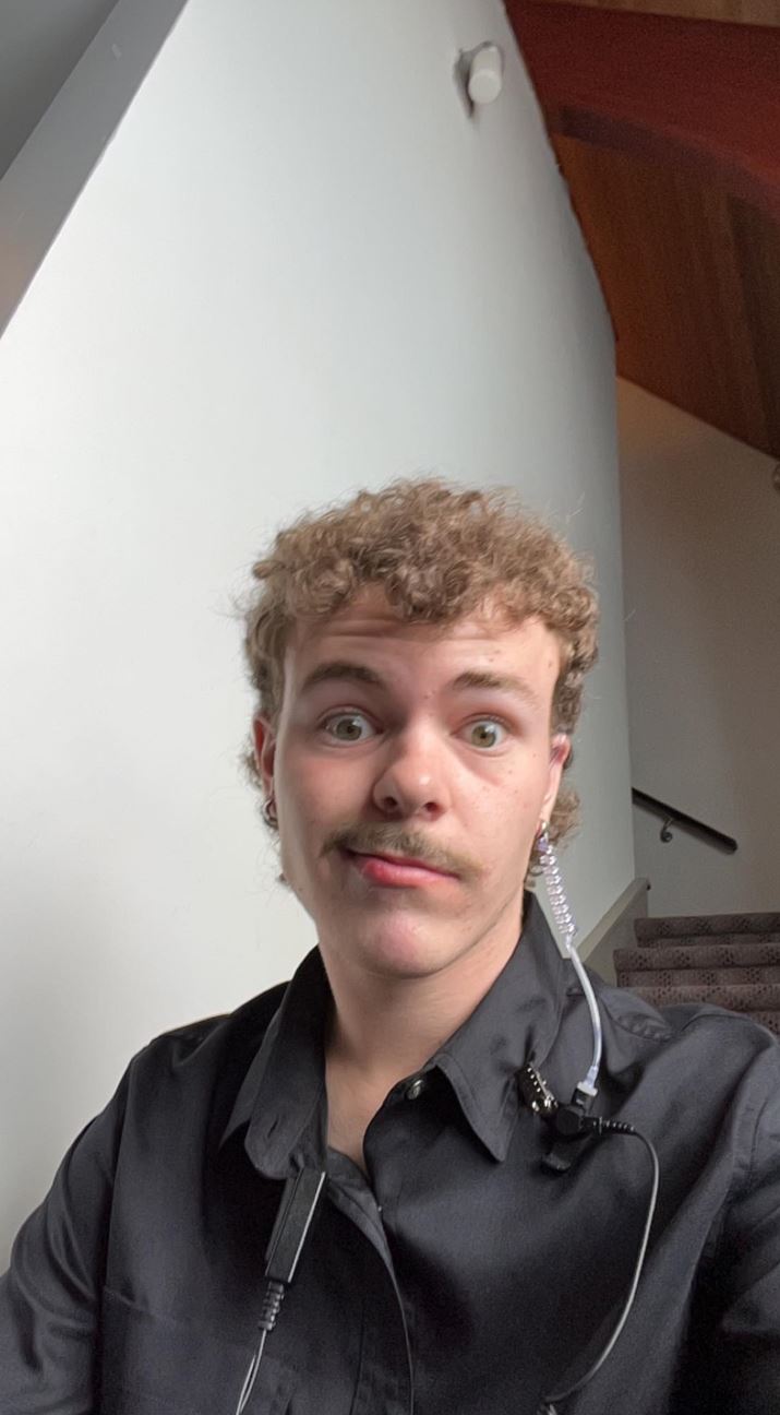 Tyler O’Dea The image shows a young man with curly blond hair and a mustache, making a playful expression. He is wearing a black shirt and has an earpiece with a coiled cord attached to it, likely indicating he is working in some capacity where communication is needed. He is indoors, near a staircase, and the background features plain white walls and wooden accents. His expression and the setting suggest a casual, light-hearted moment.