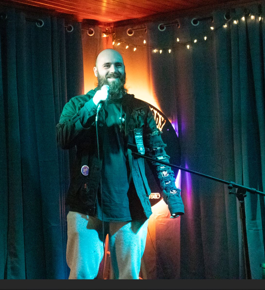 eric Subject Person: A bearded man performing stand-up comedy. Appearance Clothing: He is wearing a dark jacket with some distressed details on the sleeves and light-colored pants. Setting: The stage has dark curtains as the backdrop with string lights hanging above. There is also a microphone stand and a wooden chair visible to the side. Background Lighting: The stage is illuminated with a combination of blue and warm lights, creating a cozy atmosphere. The background includes a circular logo partially visible behind the performer. This image captures a moment from a comedy performance, emphasizing the relaxed and intimate setting of the venue.