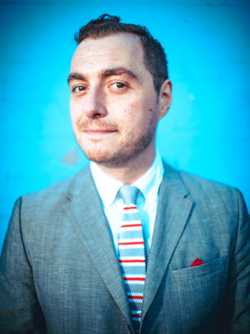 OLYMPUS DIGITAL CAMERA The image shows a man with short dark hair and a subtle smile. He is wearing a light gray suit, a white shirt, and a striped tie with colors including red and blue. He also has a red pocket square in his suit jacket pocket. The background is a bright blue wall, which contrasts with his attire and makes him stand out. His expression is confident and approachable.