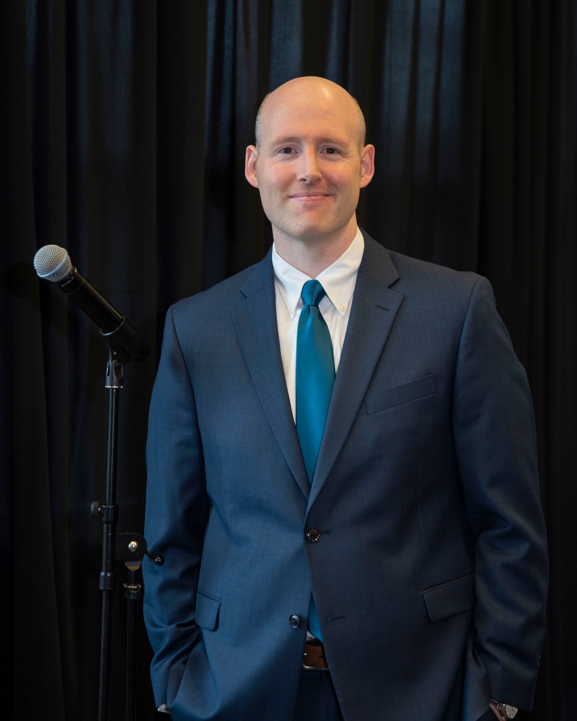 Christopher Tobin This image shows a man with a light complexion, who is bald and dressed in a formal suit. He is wearing a blue suit jacket, a white dress shirt, and a teal tie. He stands with one hand in his pocket and is smiling slightly, posing next to a microphone stand. The background features a black curtain, giving the setting a professional or event atmosphere.
