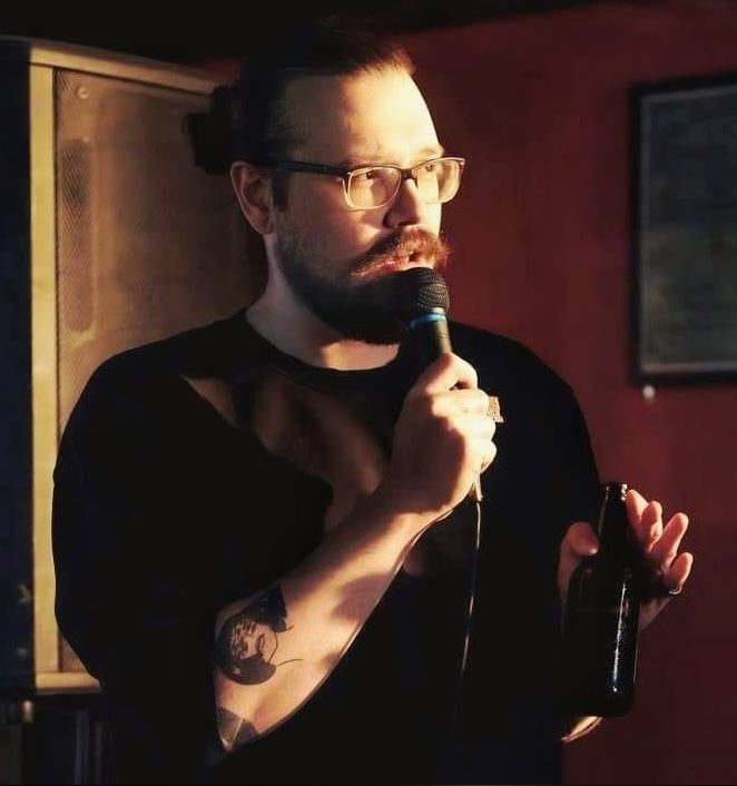 Kevin Coffey The photo features a male comedian, Kevin Coffey, on stage. He has a distinctive mustache and beard, and he is wearing glasses. His hair is pulled back into a bun. He is holding a microphone in one hand and a bottle in the other. The lighting is warm, casting a glow on his face. He is dressed in a dark shirt, and his right arm has a visible tattoo. The background is dimly lit, focusing attention on him as he performs.