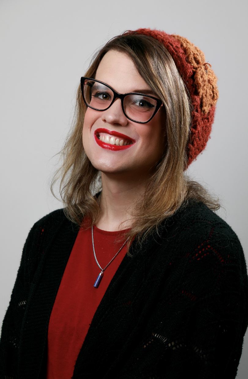 Krystal Lynn Hiller The image features a person with a cheerful expression, wearing a red top and a black cardigan. They have a beige and brown knitted hat with a decorative flower on the side. They have long, light brown hair and are wearing black-rimmed glasses. They also have a bright red lipstick and are smiling broadly. They are wearing a necklace with a blue pendant. The background is plain and light-colored, keeping the focus on the subject.