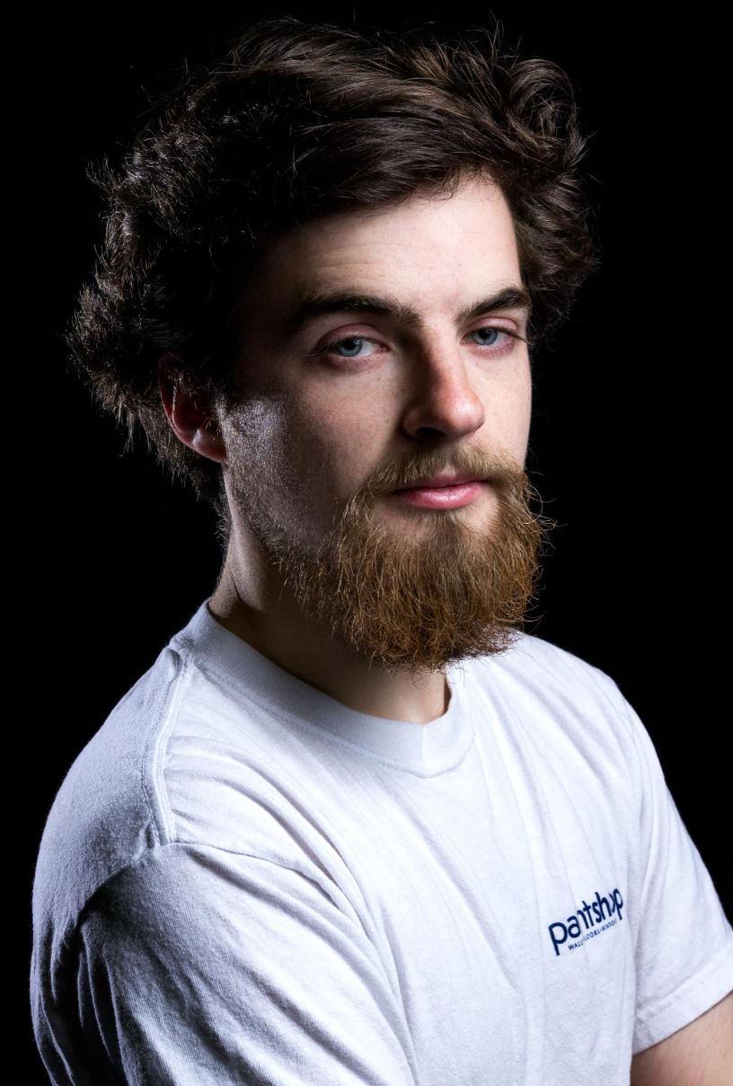 Liam Penney This image shows a young man with a light complexion, a bushy brown beard, and tousled brown hair. He is wearing a white T-shirt with a small logo on the left chest that reads "Paintshop." He has a serious expression and is looking directly at the camera. The background is black, providing a stark contrast to his light-colored clothing and skin.