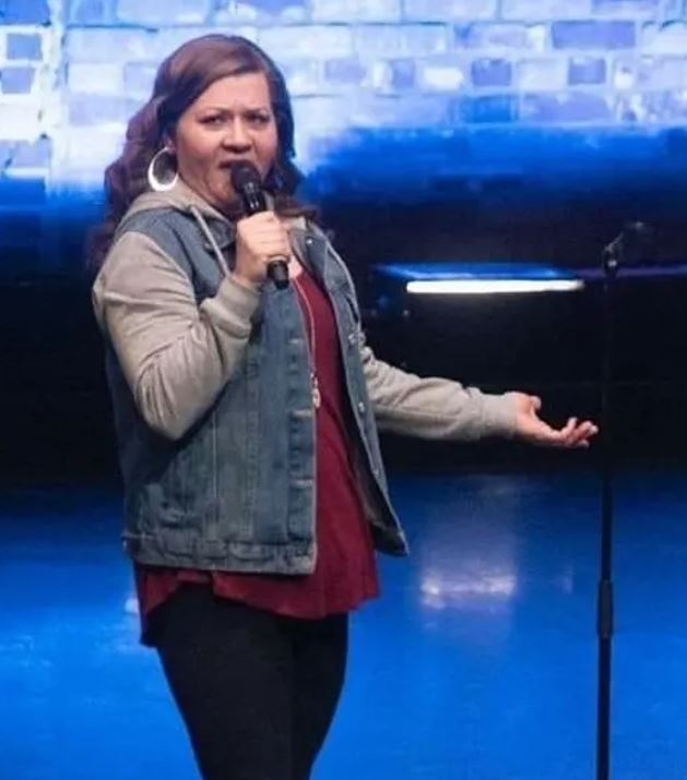 Lisa Baker The image shows Lisa Baker performing on stage. She is holding a microphone in her right hand and gesturing with her left hand. Lisa is wearing a denim vest over a maroon top and dark pants. Her expression appears animated as she engages with the audience. The background features a blue-lit wall, possibly with a brick pattern, and stage lighting.