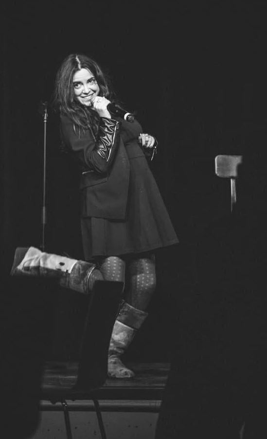 Lisa Jane The black and white photo features a female comedian on stage. She has long, wavy hair and is smiling while holding a microphone close to her mouth. She is wearing a dark jacket over a dress, patterned tights, and tall boots. One of her legs is playfully lifted behind her as she leans slightly to the side, adding a lively and engaging touch to her performance. The background is dark, drawing attention to her expressive pose and joyful expression.