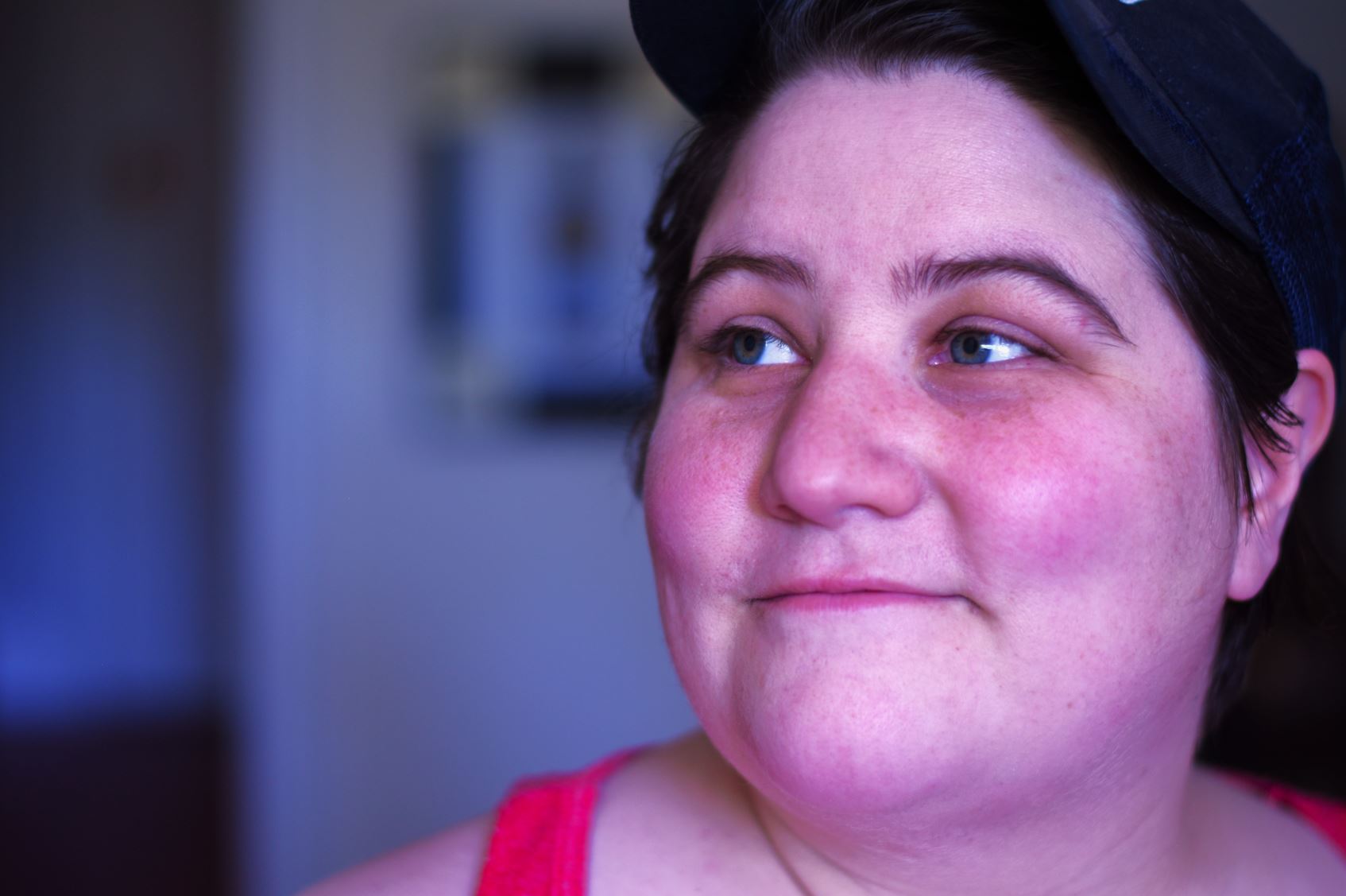 Veronica Dymond This image is a close-up portrait of a person with a light complexion. They have short, dark hair and are wearing a black baseball cap. The person is looking off to the right with a slight, serene smile. Their cheeks are slightly flushed, and they have a few freckles. They are dressed in a bright pink tank top. The background is softly blurred, putting the focus entirely on their face.