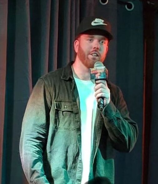 adam The image shows a man on stage holding a microphone, appearing to be mid-performance. He is wearing a black Nike cap, a dark denim jacket over a white t-shirt, and dark jeans. The setting seems to be a small, intimate venue with a dark curtain as the backdrop, creating a focused atmosphere on the performer. The man has a reddish beard and is speaking into the microphone, likely engaging with the audience.