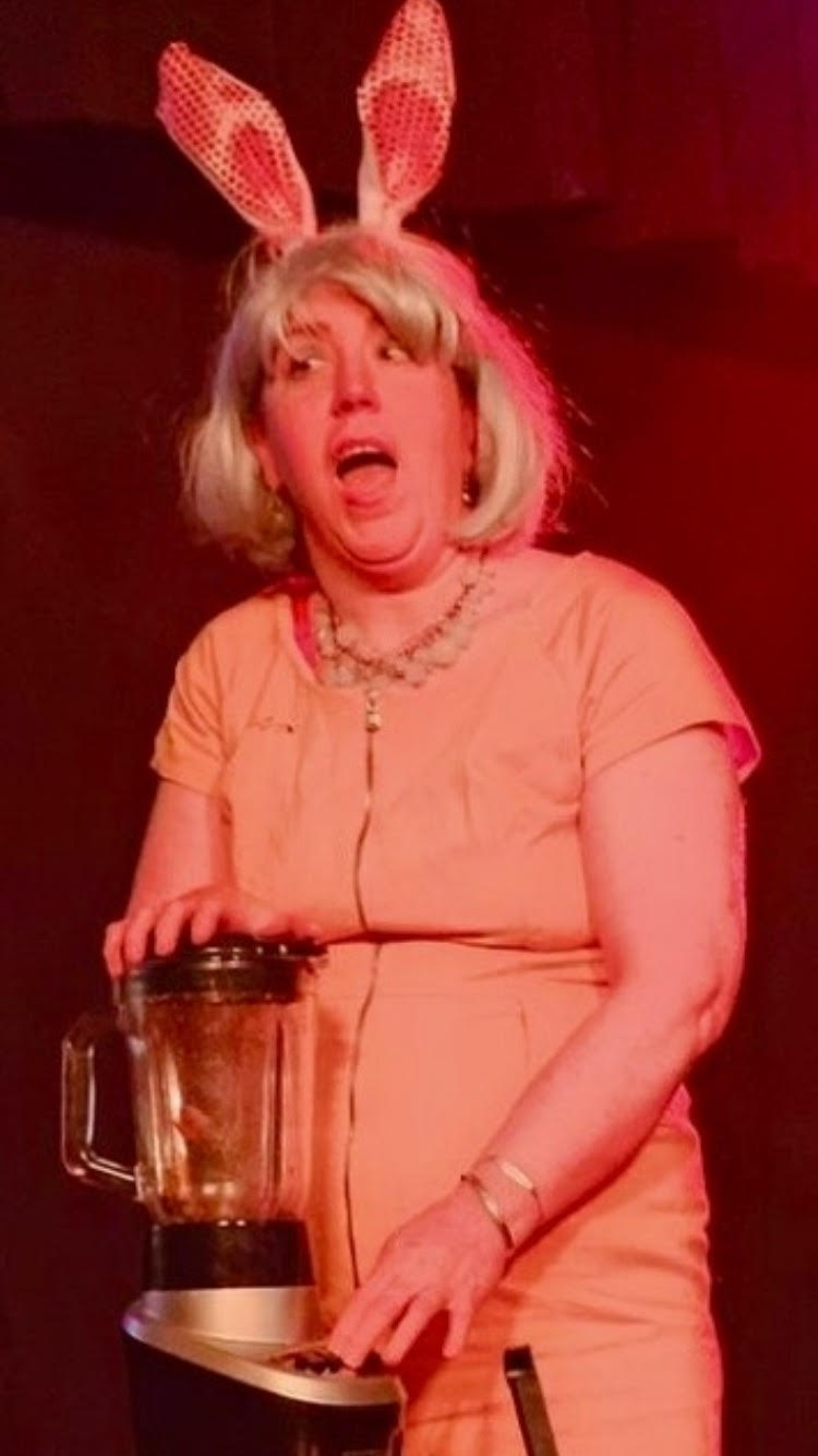 Ellen Reid The image shows a performer mid-act, dressed in a peach outfit with a bold necklace and bunny ears, holding onto a blender. The lighting creates a dramatic and comedic atmosphere, likely capturing a humorous moment from a live show. The person’s expression adds to the lively and engaging nature of the performance, suggesting a playful or theatrical context.