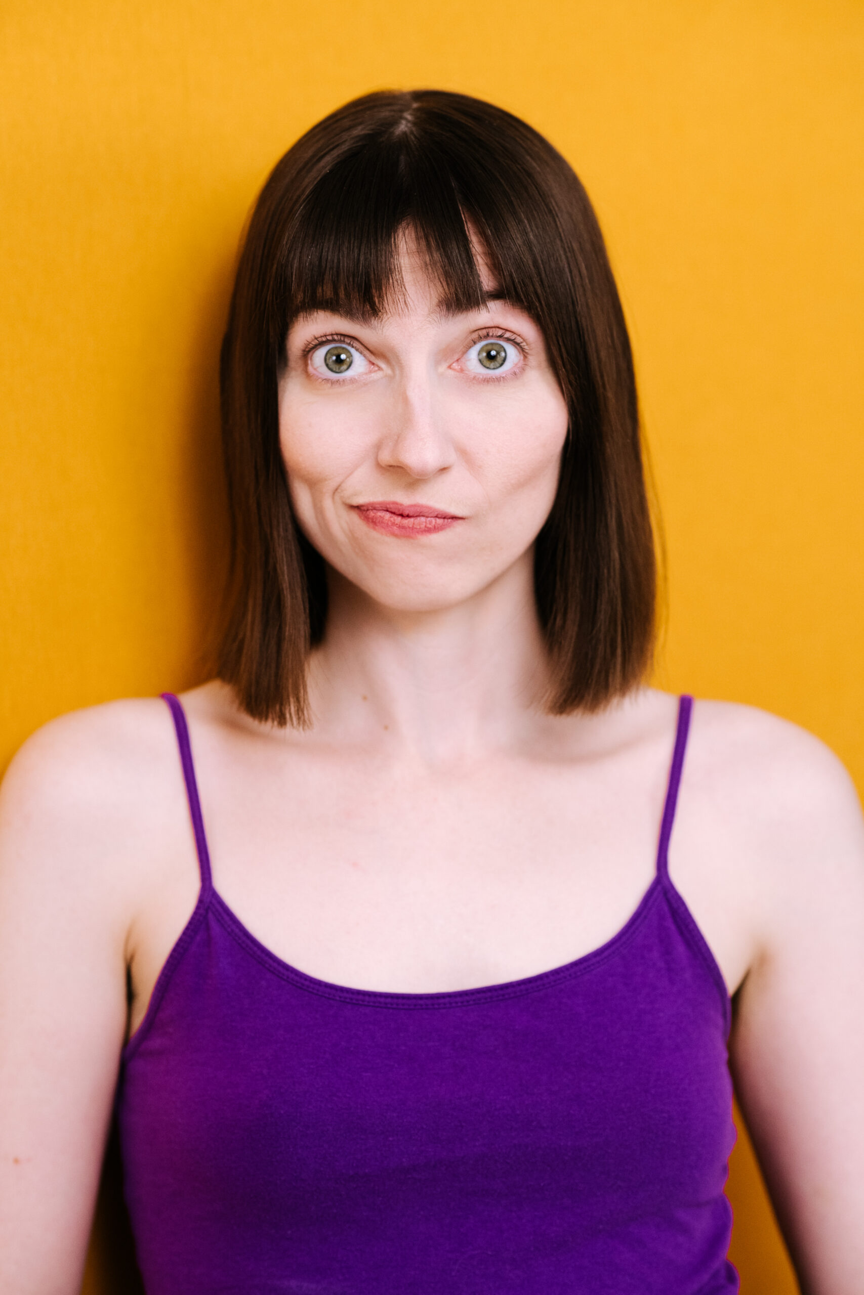 JanaGillis-LVIMAGERY-3 This is a portrait of a young woman with straight brown hair cut in a neat, shoulder-length bob and bangs. She has strikingly large, light green eyes and a small, subtle smile. Her expression is friendly and open, with a slight air of curiosity or amusement. She is wearing a vibrant purple sleeveless top, and the background is a solid, cheerful yellow, which creates a striking contrast with her outfit and hair, making her features stand out prominently. The overall tone of the photo is bright and inviting.
