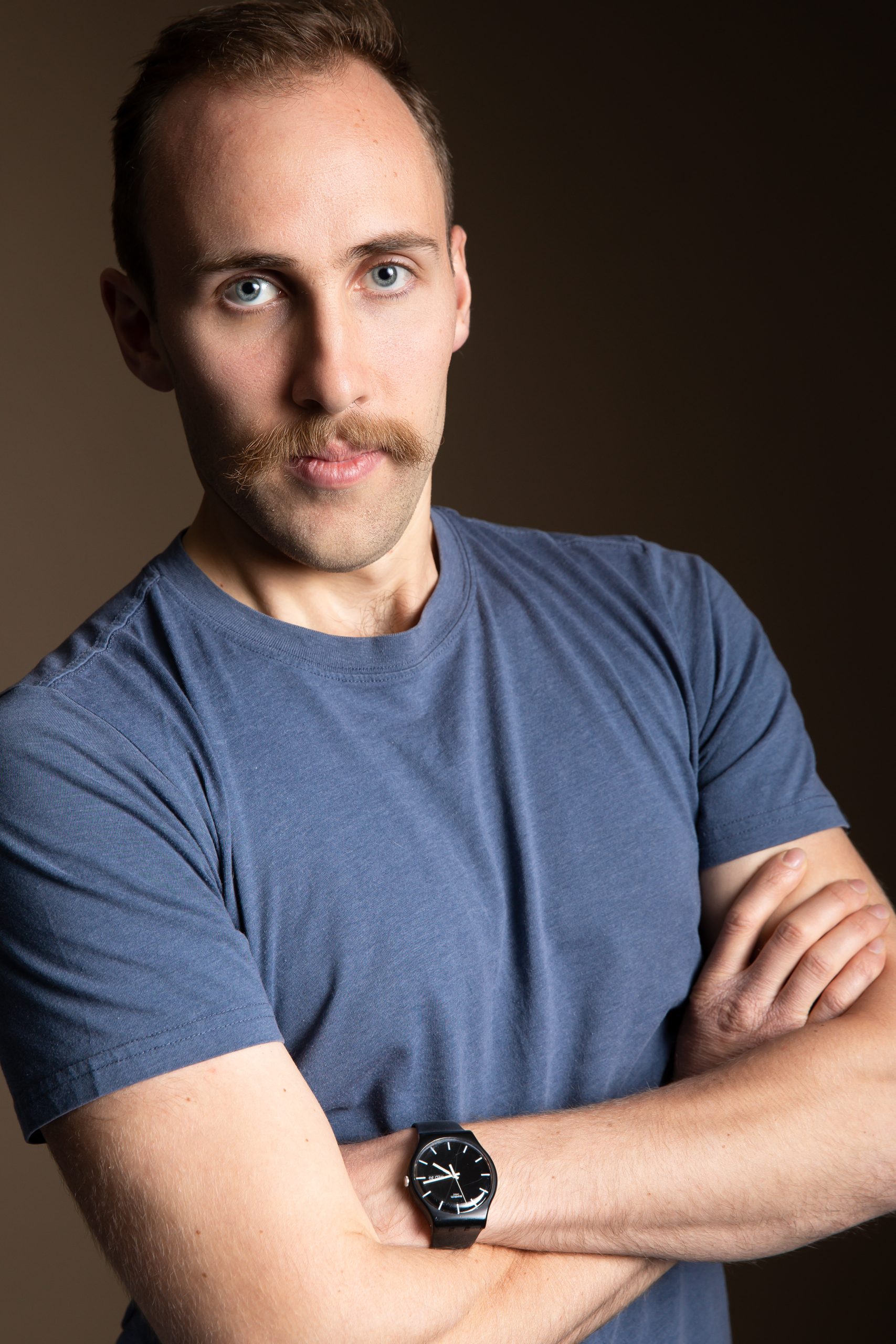 645BD13B-7DBE-4790-A676-70BCF6F1B764 This is a professional portrait of a man with light skin, blue eyes, and a distinctive mustache. He has a confident expression, with his arms crossed, and is wearing a fitted blue T-shirt and a black wristwatch with a sleek design. The background is a simple, neutral-toned setting, giving the photo a polished, studio-like appearance. The lighting is soft and even, highlighting his facial features and giving the image a clean, professional feel.